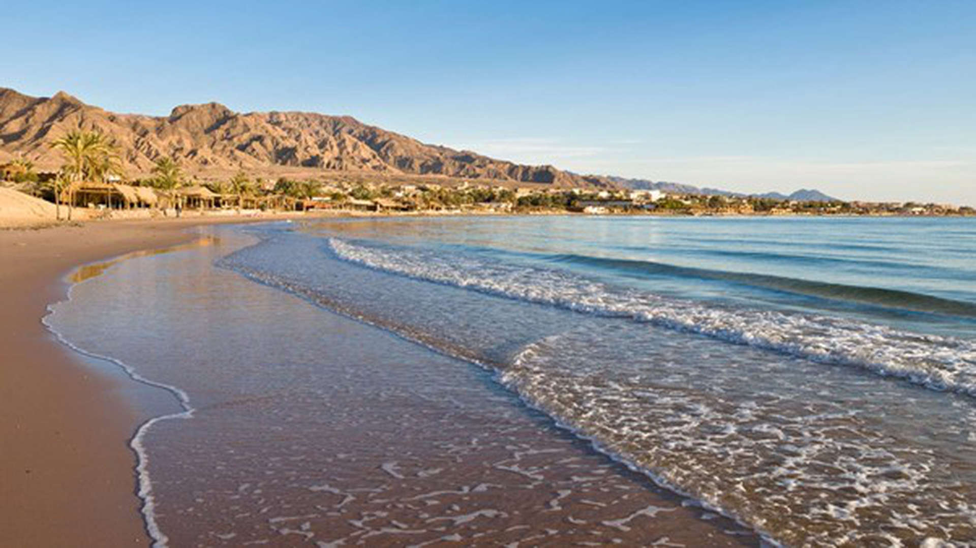 Dahab Strand in Ägyptens Sinai-Halbinsel. Ein goldener Strand mit leichten Wellen aus klarem Wasser und Bergen am Horizont.