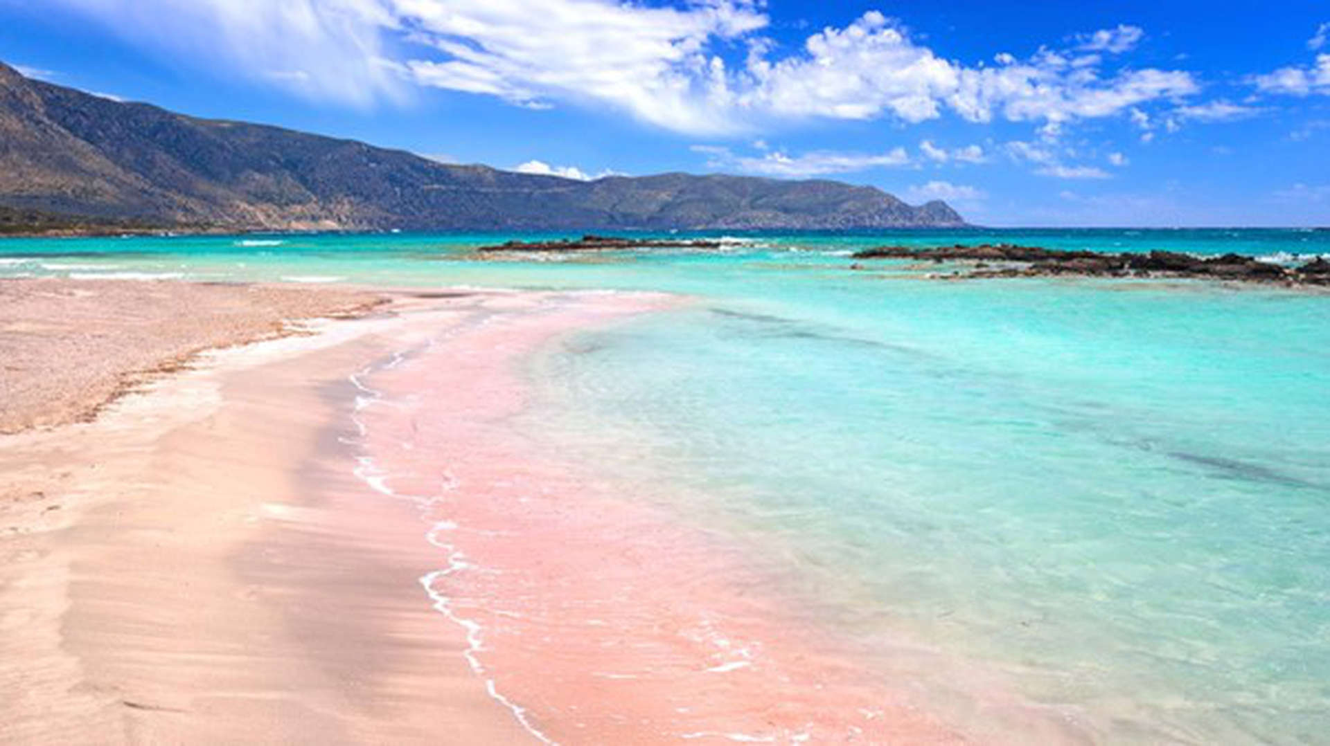 Elafinisi Strand auf der Insel Kreta in Griechenland. Ein rosafarbener Strand der von Türkisem Wasser eingesäumt wird, mit Bergen im Hintergrund.
