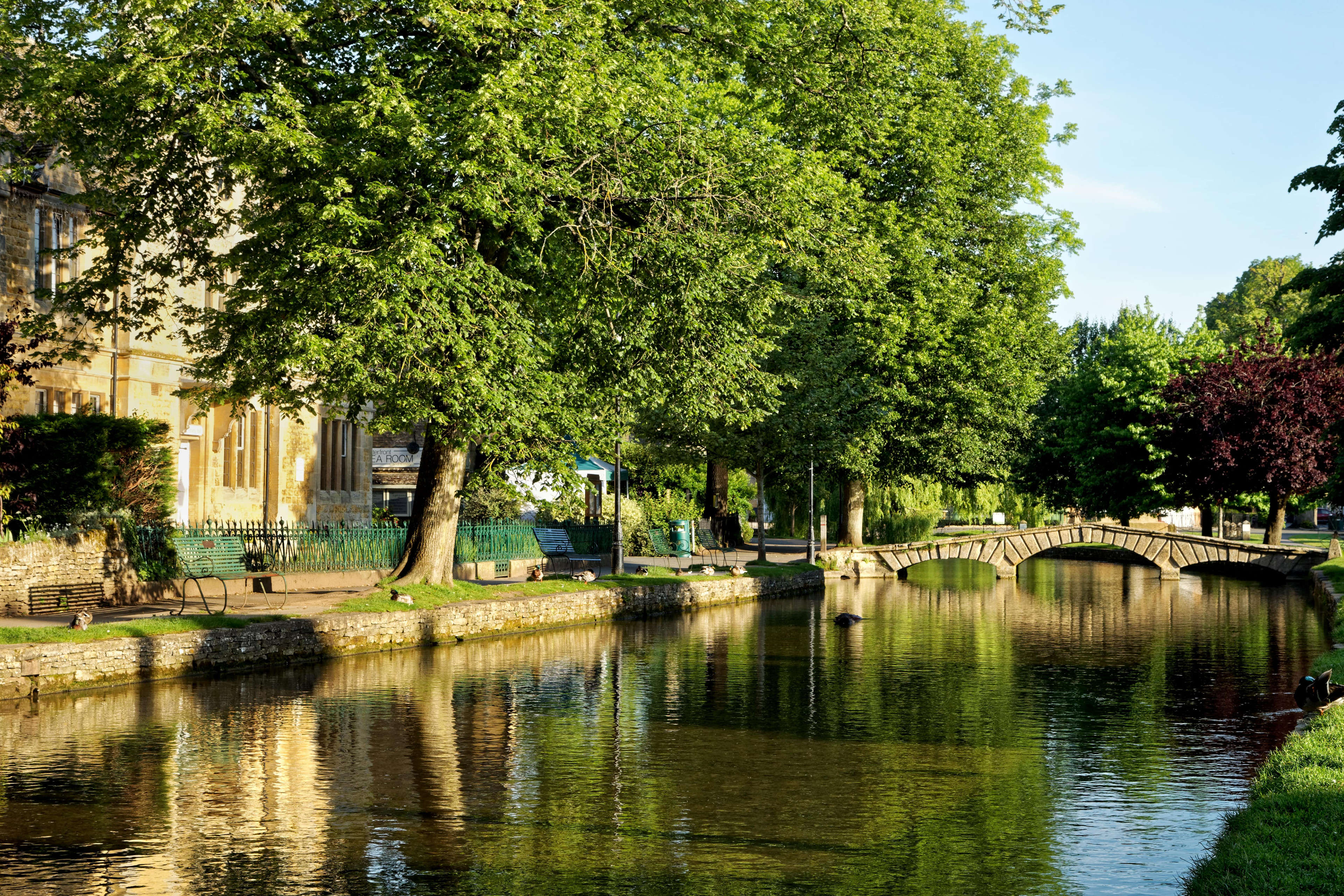 View over Windrush river with summer trees, footbridge and reflections in Bourton-on-the-Water in the Cotswolds, Gloucestershire, England