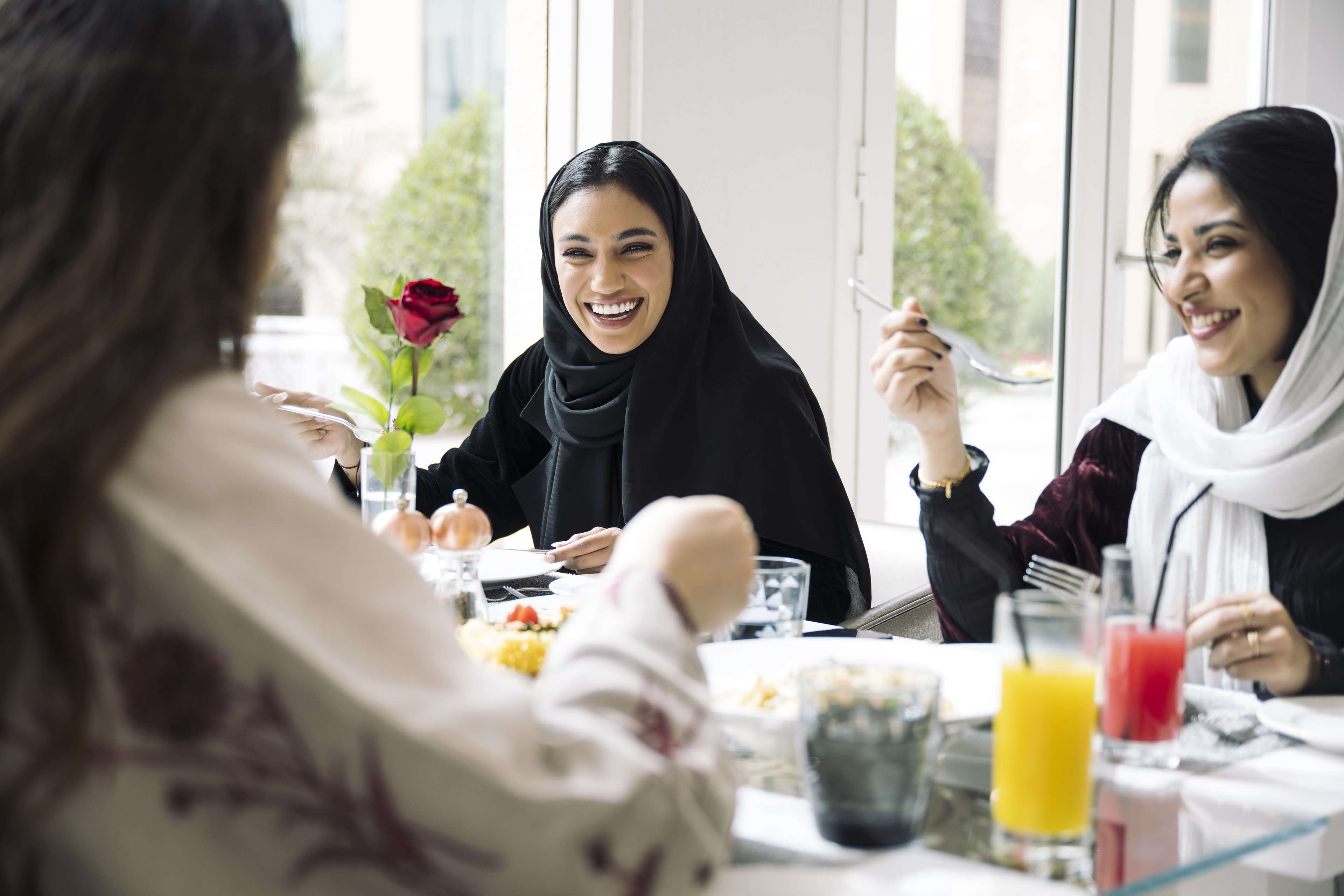 Young Saudi women enjoying lunch in Riyadh restaurant Waist-up view of Middle Eastern friends in abayas and headscarves talking and laughing as they eat a light midday meal at sunny window table